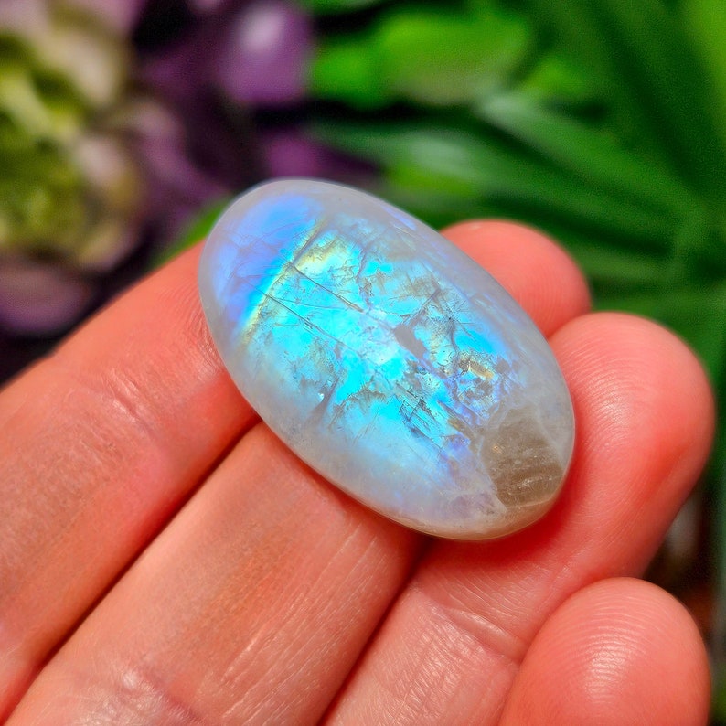 May include: A white oval moonstone cabochon with iridescent blue and green flashes. The stone is smooth and polished.
