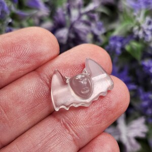 May include: A translucent, light pink bat-shaped charm with a detailed face carved into the center. The charm has pointed ears and wings, and is held in a person's hand. The background is blurred with purple flowers.