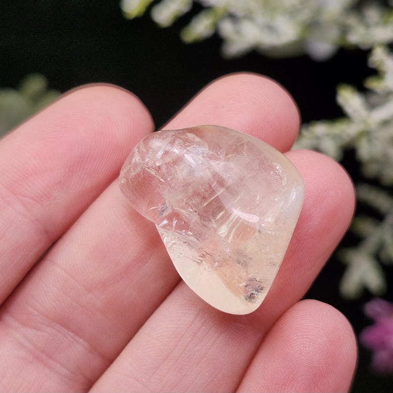 May include: A polished, translucent, light yellow-colored gemstone. The stone has a smooth, rounded shape and appears to be held in a hand. The background is blurred, with hints of green and white foliage.