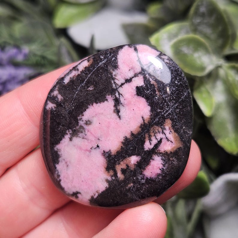 May include: A polished, rounded rhodonite stone featuring a smooth surface with a distinct pattern of opaque pink and deep black mineral inclusions. The stone has a glossy finish that reflects light, highlighting the contrast between the colors.