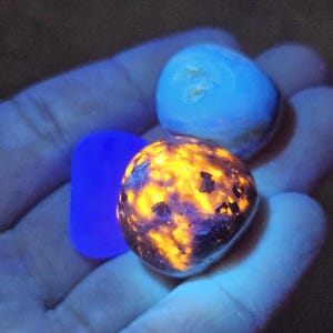 May include: A hand holds three glowing stones. One is a vibrant orange and black, another is a light blue, and the third is a deep blue rectangle. The stones appear to be illuminated by a black light.