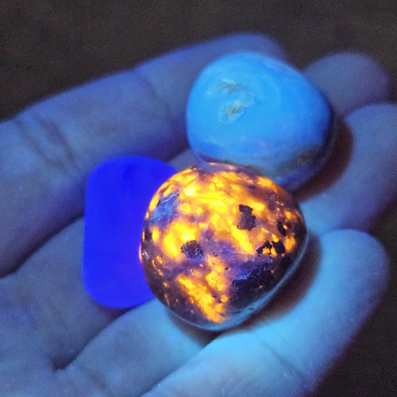 May include: A hand holds three glowing stones. One is a vibrant orange and black, another is a light blue, and the third is a deep blue rectangle. The stones appear to be illuminated by a black light.