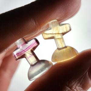 May include: Two small, translucent cross-shaped carvings. One is a mix of purple and white, the other is yellow. The crosses are held between fingers, showcasing their detailed design and polished finish. The base of each cross is rounded.