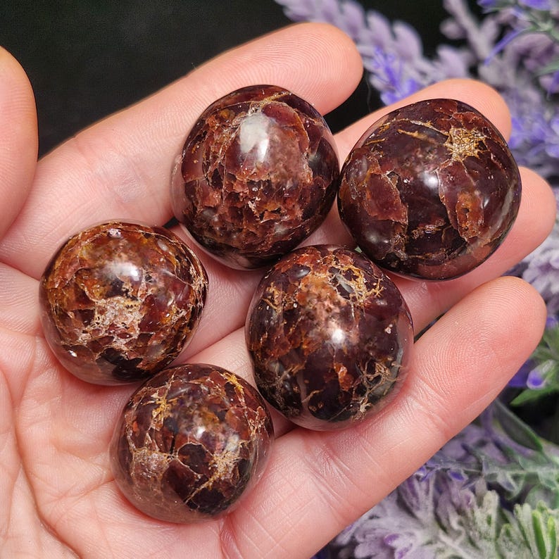 May include: Five polished, round gemstones in varying shades of deep red and brown are held in a hand. The stones have a glossy finish and a natural, textured appearance. The gemstones are a natural product.