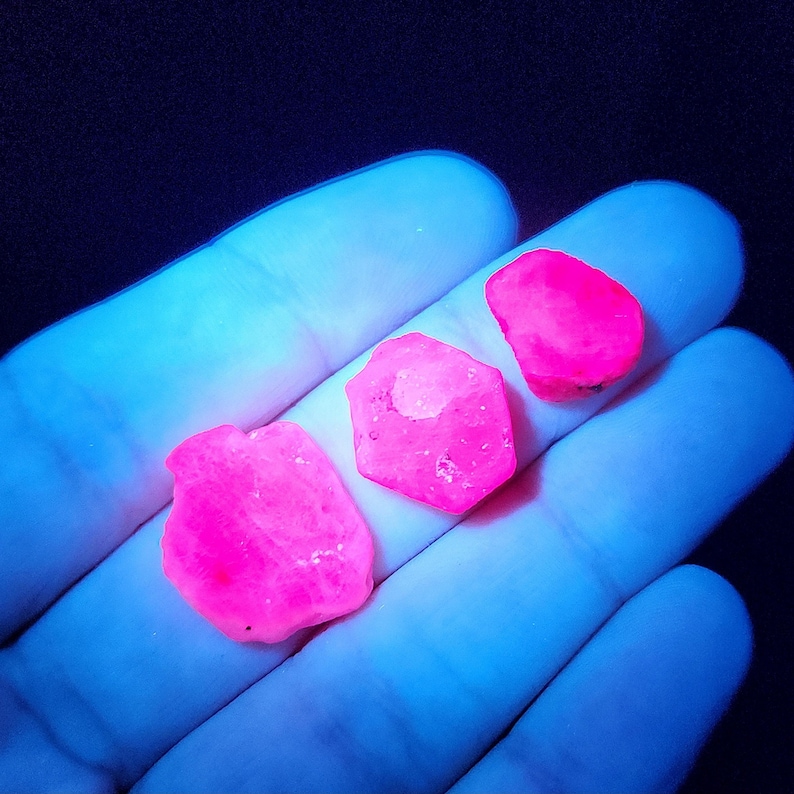 May include: Three pink gemstones, two are hexagonal and one is oval, under a UV light.
