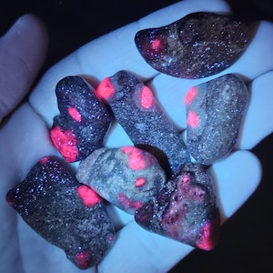 May include: A collection of six dark gray stones with bright pink accents. The stones are smooth and have a variety of shapes and sizes.