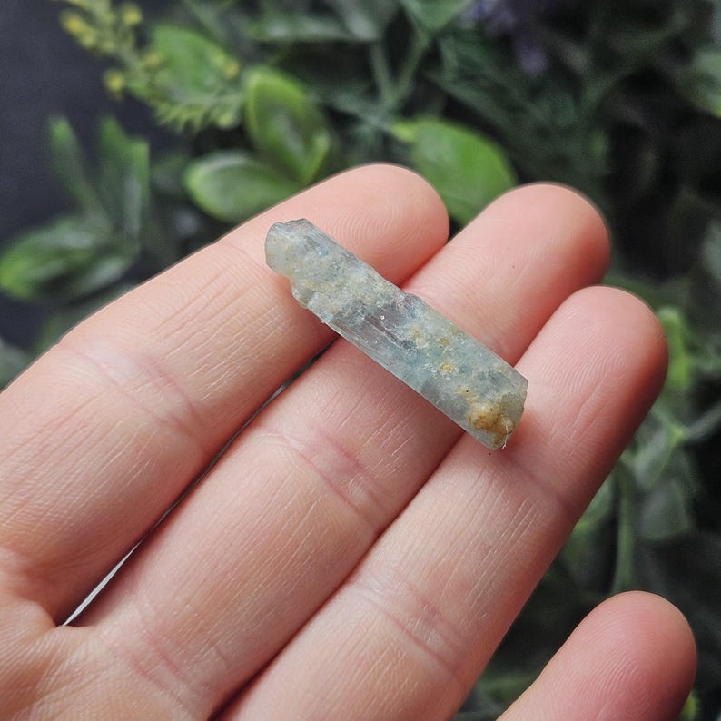 Puede incluir: Un cristal de aguamarina en bruto, de color azul claro, con una textura ligeramente rugosa. El cristal se sostiene en una mano, mostrando su forma natural y variaciones de color. La piedra tiene un aspecto transl&uacute;cido.