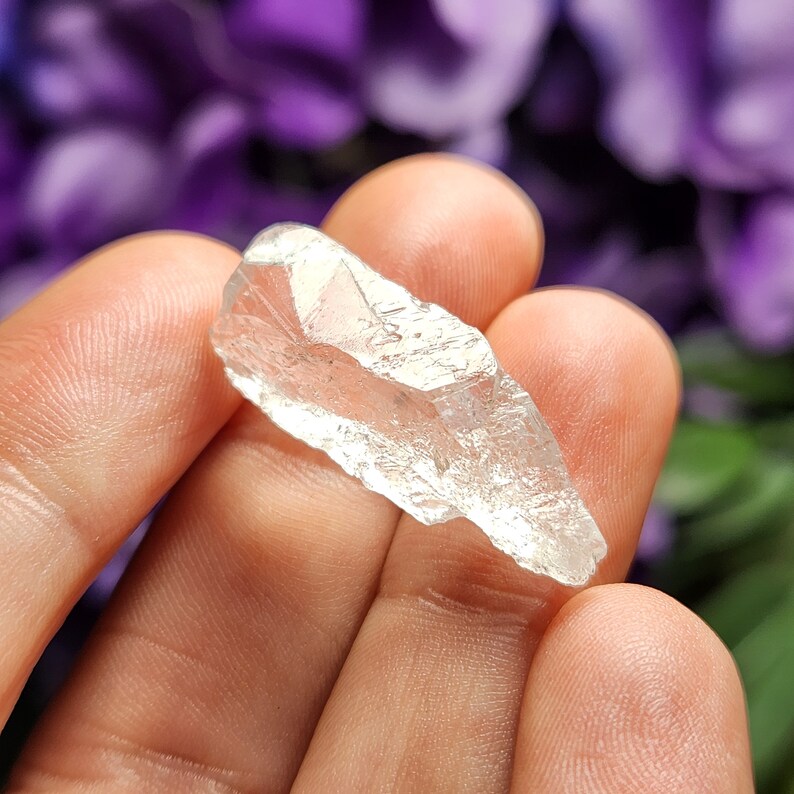 May include: A clear quartz crystal point with a rough, natural texture. The crystal is held in a hand, showcasing its size and shape.