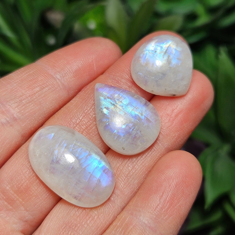 May include: Three iridescent white moonstone cabochons in teardrop shapes. The stones are translucent and have a blue and purple sheen.