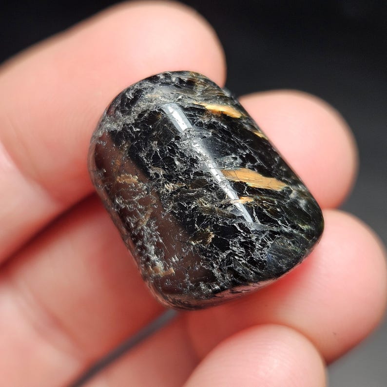 May include: A polished black gemstone with gold-colored inclusions. The stone has a smooth, rounded shape and a glossy surface. The gemstone is held in a person's hand. The stone is approximately 1.5 inches long.