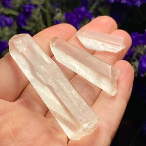May include: Three clear quartz crystal points of varying sizes, held in a hand. The crystals are smooth and translucent, with a slight shimmer.