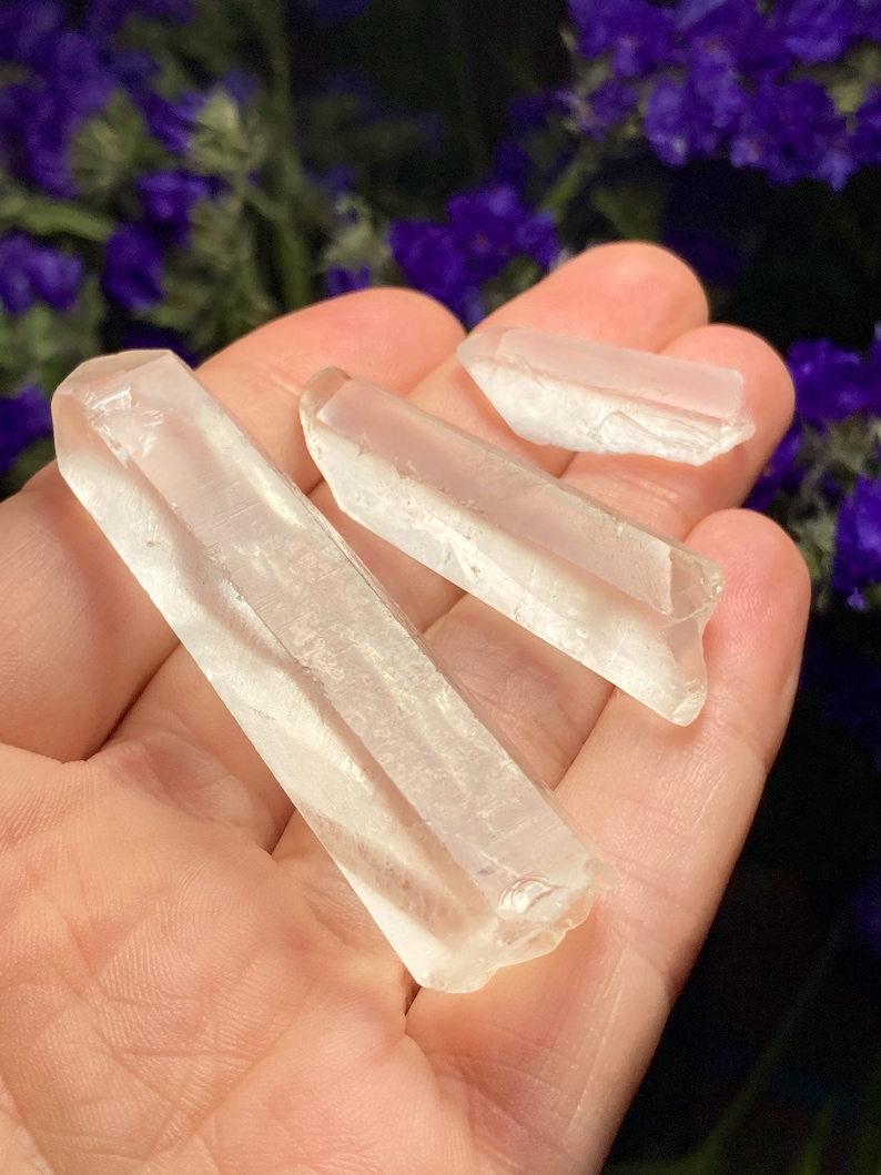 May include: Three clear quartz crystal points of varying sizes, held in a hand. The crystals are smooth and translucent, with a slight shimmer.