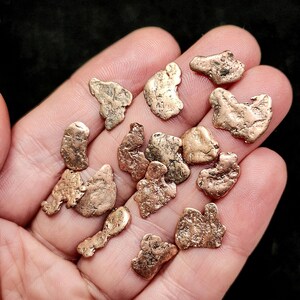May include: A collection of irregularly shaped, metallic gold nuggets. The nuggets vary in size and have a textured, rough surface. The color is a warm, golden brown. The nuggets are held in an open hand against a dark background.