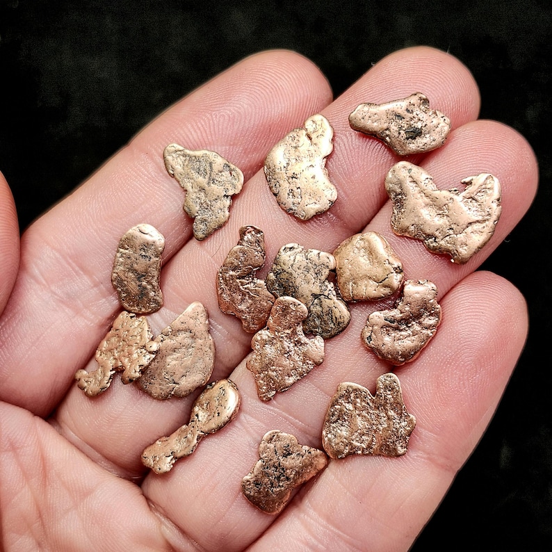 May include: A collection of irregularly shaped, metallic gold nuggets. The nuggets vary in size and have a textured, rough surface. The color is a warm, golden brown. The nuggets are held in an open hand against a dark background.