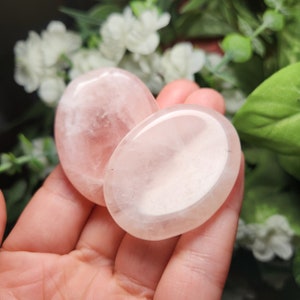 May include: Two smooth, oval-shaped rose quartz stones, one slightly larger than the other, with a translucent, pale pink color. The stones are held in a hand, with the larger stone on top of the smaller one.