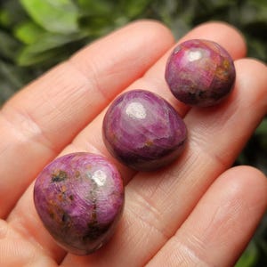 May include: Three polished, oval-shaped gemstones in varying shades of purple and pink are displayed. The stones have a smooth, reflective surface with natural inclusions and variations in color. The gemstones are held in an open hand.