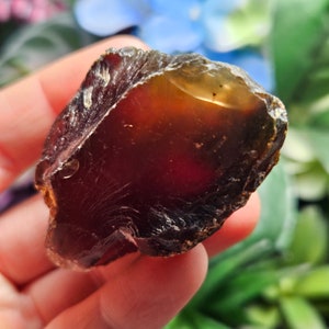 May include: A large, dark red, amber-colored, rough-cut gemstone with a smooth, polished surface. The gemstone has a translucent, glassy appearance and is held in a hand.