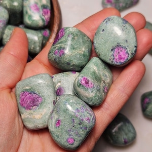May include: A collection of tumbled ruby in zoisite stones. The stones are a mottled green color with vibrant pink ruby inclusions. The stones are smooth and polished, with a natural, organic shape. The stones are held in a hand.