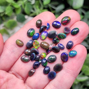 May include: A collection of oval-shaped black opals with iridescent flashes of color, including green, blue, and red. The gemstones are displayed in a hand, showcasing their polished surfaces and natural variations. The opals vary in size and shape.