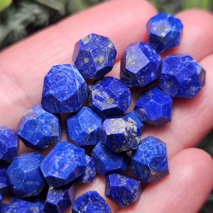 May include: A collection of small, faceted lapis lazuli beads featuring a deep blue color with natural gold-colored pyrite inclusions. Each bead has a multi-sided geometric shape with sharp edges and a polished finish, displaying unique patterns.