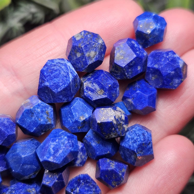 May include: A collection of small, faceted lapis lazuli beads featuring a deep blue color with natural gold-colored pyrite inclusions. Each bead has a multi-sided geometric shape with sharp edges and a polished finish, displaying unique patterns.