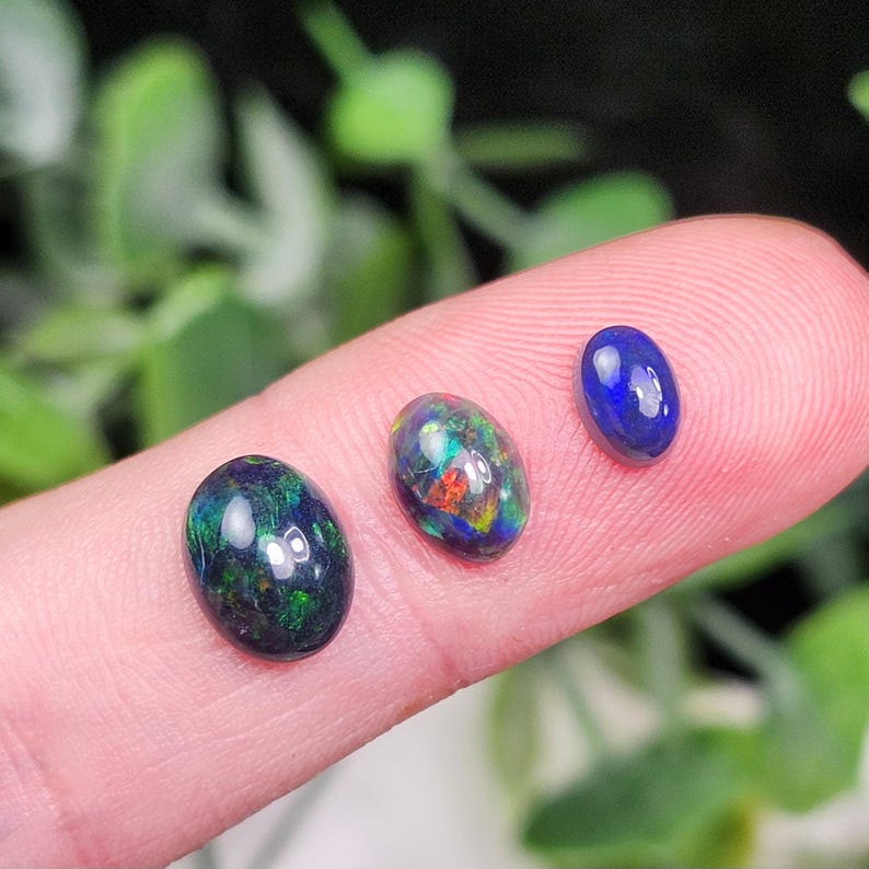 May include: Three oval-shaped opals in shades of dark blue, green, and a mix of colors. The opals are displayed on a finger, with a blurred green background. The opals have a polished, glossy finish.