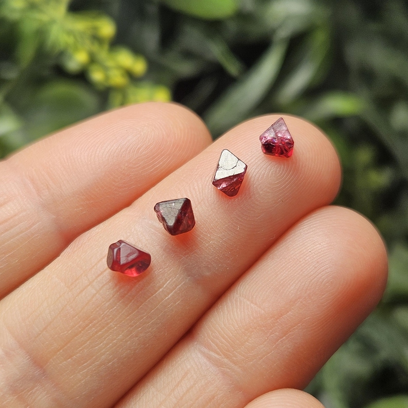 May include: Four red, faceted, natural, spinel crystals of varying sizes. The crystals are displayed on a hand.