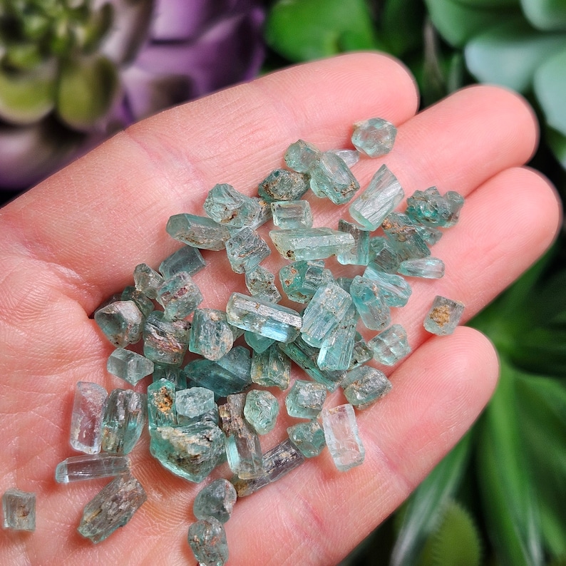 May include: A collection of small, green, translucent, and rough-textured emerald crystals. The crystals are in various shapes and sizes, and some have a slightly metallic sheen.