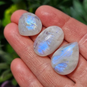 May include: Three iridescent moonstone cabochons in a teardrop and oval shape. The stones are a milky white color with a blue iridescent sheen.