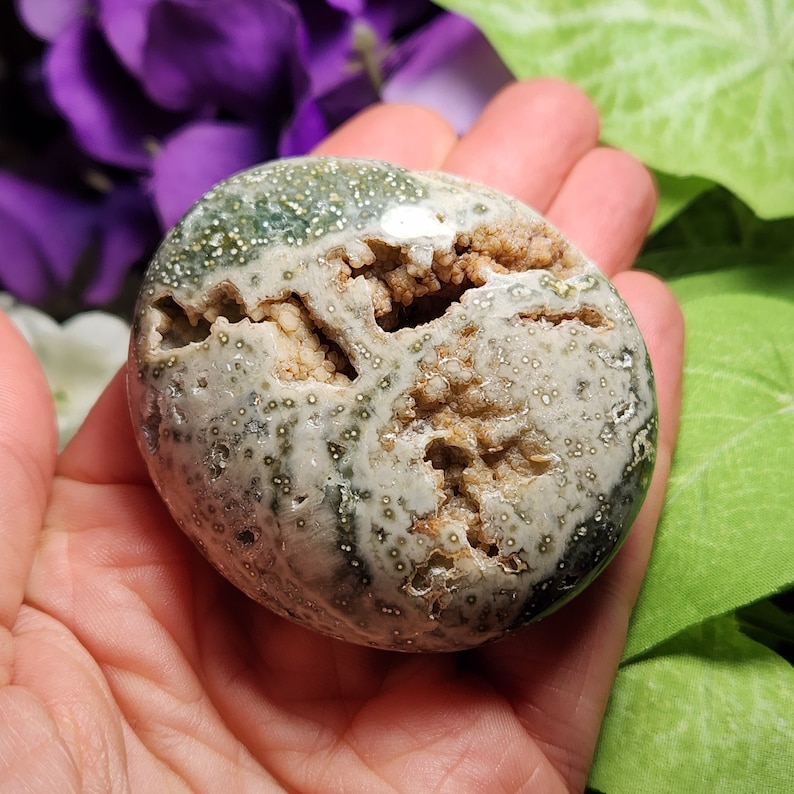 May include: A polished, oval-shaped gemstone with a geode-like interior. The exterior is a mottled mix of green, white, and gray, speckled with small, dark spots. The center reveals a cavity filled with crystalline formations.