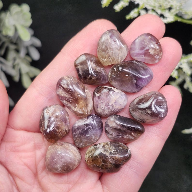 May include: A handful of polished amethyst stones, each with a unique blend of purple, brown, and clear hues. The stones vary in size and shape, with a smooth, rounded appearance. The gemstones are held in an open hand, showcasing their natural beauty.