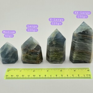May include: Four fluorite crystal towers of varying sizes, labelled Medium (60g+), Large (100g+), X-Large (125g+), and XX-Large (150g+). The crystals are shades of green, blue, and brown. A yellow ruler is at the bottom.