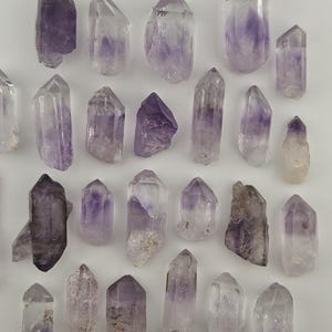 May include: An overhead shot displays a collection of amethyst crystals. The crystals vary in size and shape, with a range of purple hues. Some are translucent, while others are more opaque, showcasing natural formations and textures. The crystals are arranged on a white surface.