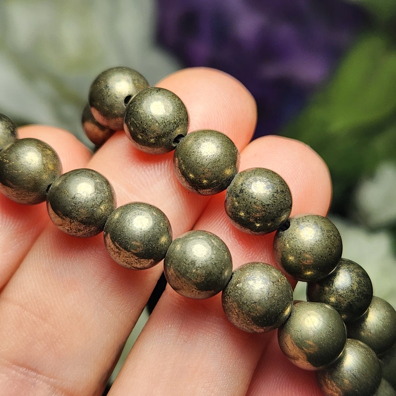 May include: A close-up of a pyrite bracelet. The bracelet features multiple spherical, metallic-looking beads in shades of gold and grey. The beads are strung together on a black cord. The background is blurred, with hints of green and purple.