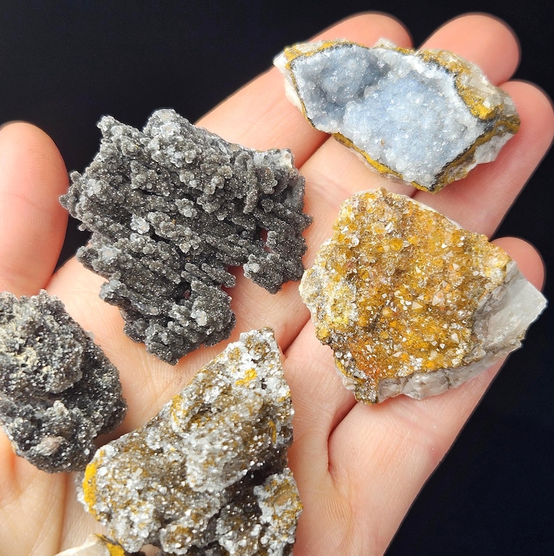 May include: A collection of raw mineral specimens, including a piece with a light blue interior, and others with dark gray and golden-brown hues. The minerals are displayed against a dark background, showcasing their natural textures and colors.