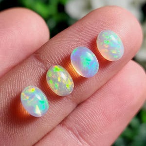 May include: Four oval-shaped cabochon opals arranged in a row. Each gemstone features a translucent white base with vibrant flashes of green, yellow, and blue fire. The stones are smooth, polished, and display a distinct play-of-color effect.