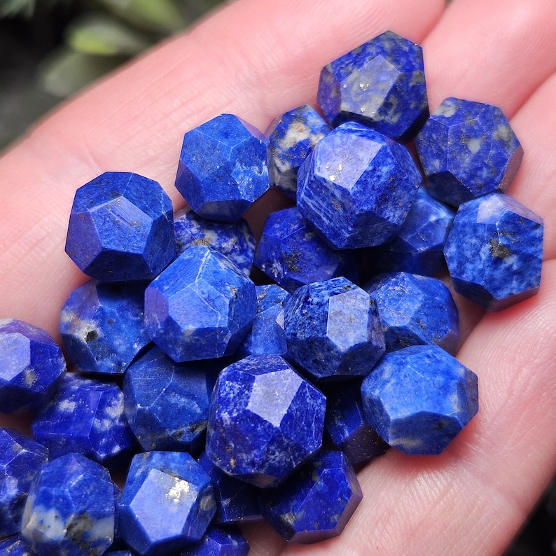 May include: A collection of small, faceted lapis lazuli gemstones. Each stone features a deep blue color with natural white and gold-colored mineral inclusions. The beads are cut into a multi-faceted, geometric shape with sharp edges and flat surfaces.