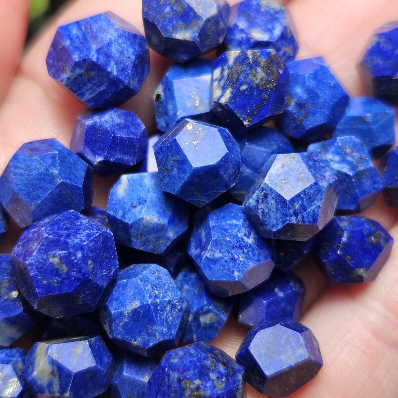 May include: A collection of small, faceted lapis lazuli gemstones with a deep blue color. Each stone features multiple flat, polished surfaces and visible flecks of gold-colored pyrite inclusions scattered throughout the intense blue mineral.