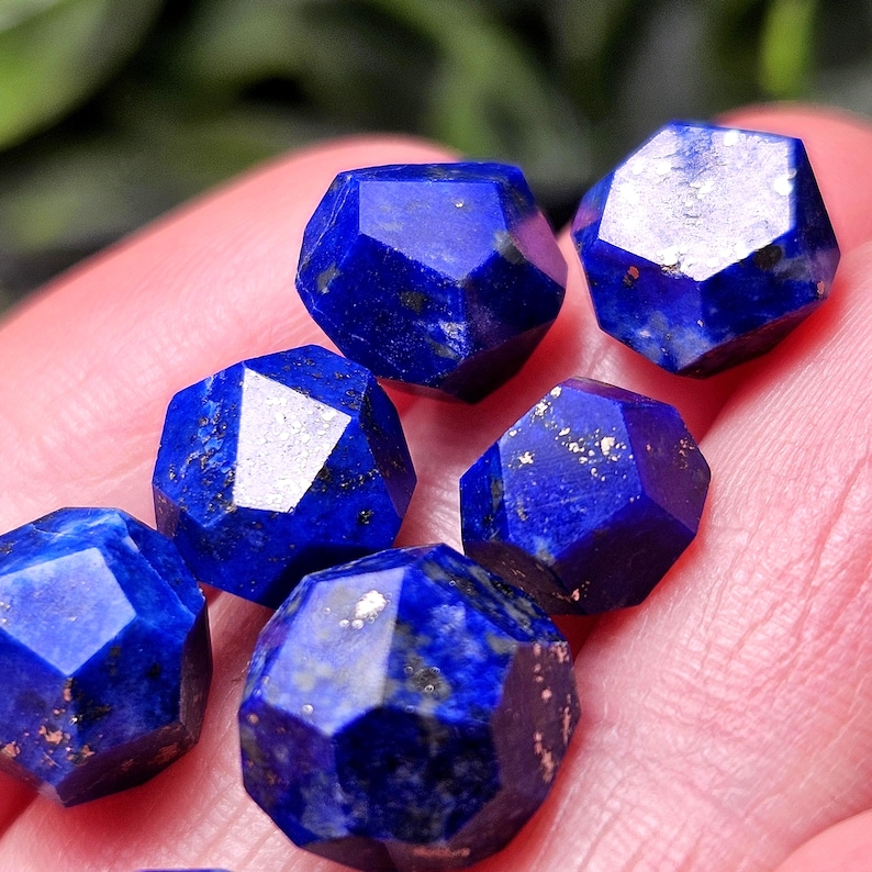 May include: A collection of six faceted lapis lazuli gemstones, each displaying a deep royal blue color with natural golden pyrite inclusions and dark mineral flecks, arranged in a cluster with geometric, multi-sided cut surfaces.