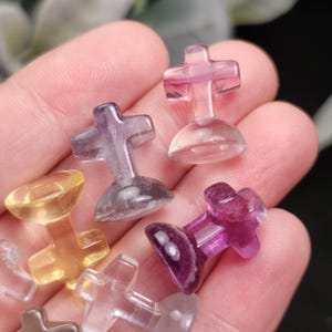 May include: A collection of small, translucent cross-shaped figurines in various colors, including purple, pink, yellow, and clear. Each cross is mounted on a rounded base. The figurines are held in a hand, showcasing their polished appearance.