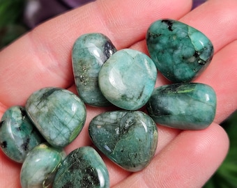 May include: A collection of tumbled emerald gemstones, each with a unique green hue and natural inclusions. The stones are smooth and polished, with a variety of shapes and sizes.
