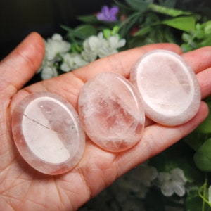 May include: Three oval-shaped rose quartz palm stones. The stones are a light pink color with white veins running through them. The stones are smooth and polished.