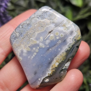 Puede incluir: Una piedra de jaspe oce&aacute;nico pulida y lisa que presenta un patr&oacute;n distintivo de orbes circulares en tonos azul suave, gris, blanco y amarillo p&aacute;lido. La superficie muestra un acabado brillante con inclusiones minerales naturales y variaciones.