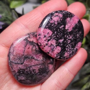 May include: Two polished, circular rhodonite stones featuring a mottled pattern of vibrant pink and deep black mineral inclusions. The stones have a smooth, glossy surface texture and irregular, organic veining throughout their rounded shapes.