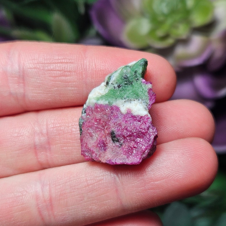 Pu&ograve; includere: Una gemma grezza multicolore. La pietra presenta una base magenta intenso, con bande di bianco e verde smeraldo. Piccole inclusioni nere sono visibili in tutta la pietra. La gemma &egrave; tenuta in mano.