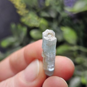 Puede incluir: Un cristal de aguamarina azul claro, sostenido entre los dedos, es el punto focal de esta imagen. El cristal tiene una textura rugosa y natural y una calidad transl&uacute;cida. El fondo est&aacute; borroso, con follaje verde.