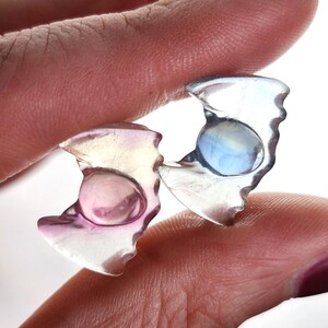 May include: Two translucent, irregularly shaped earrings with a central, rounded, iridescent stone. One earring has a pink hue, the other a blue hue. The earrings are held between fingers against a white background.