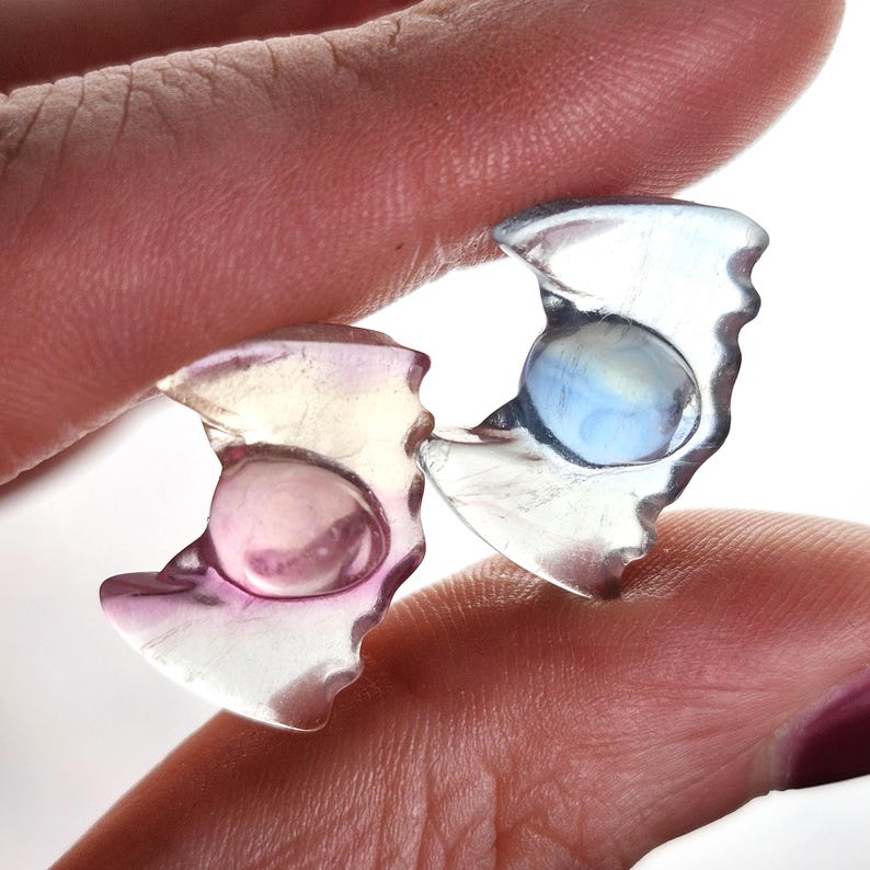 May include: Two translucent, irregularly shaped earrings with a central, rounded, iridescent stone. One earring has a pink hue, the other a blue hue. The earrings are held between fingers against a white background.