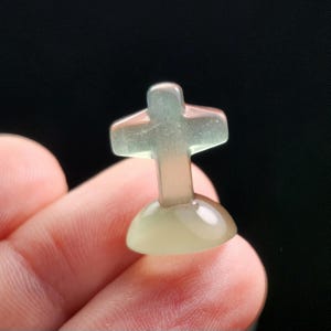 May include: A small, translucent cross figurine with a rounded base. The cross is pale green and pink, with a smooth, polished surface. The figurine is held between fingers against a dark background.