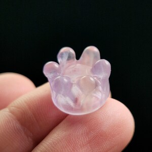 May include: A small, translucent, light purple crystal carving in the shape of a flower or hand. The object has rounded petals or fingers and a smooth, polished surface. The background is a solid black.